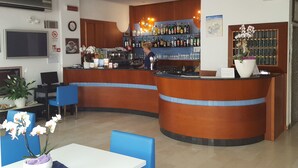 Zona de bar con mostrador, estanterías llenas de botellas y una persona trabajando detrás. Se ven sillas y una mesa en primer plano.