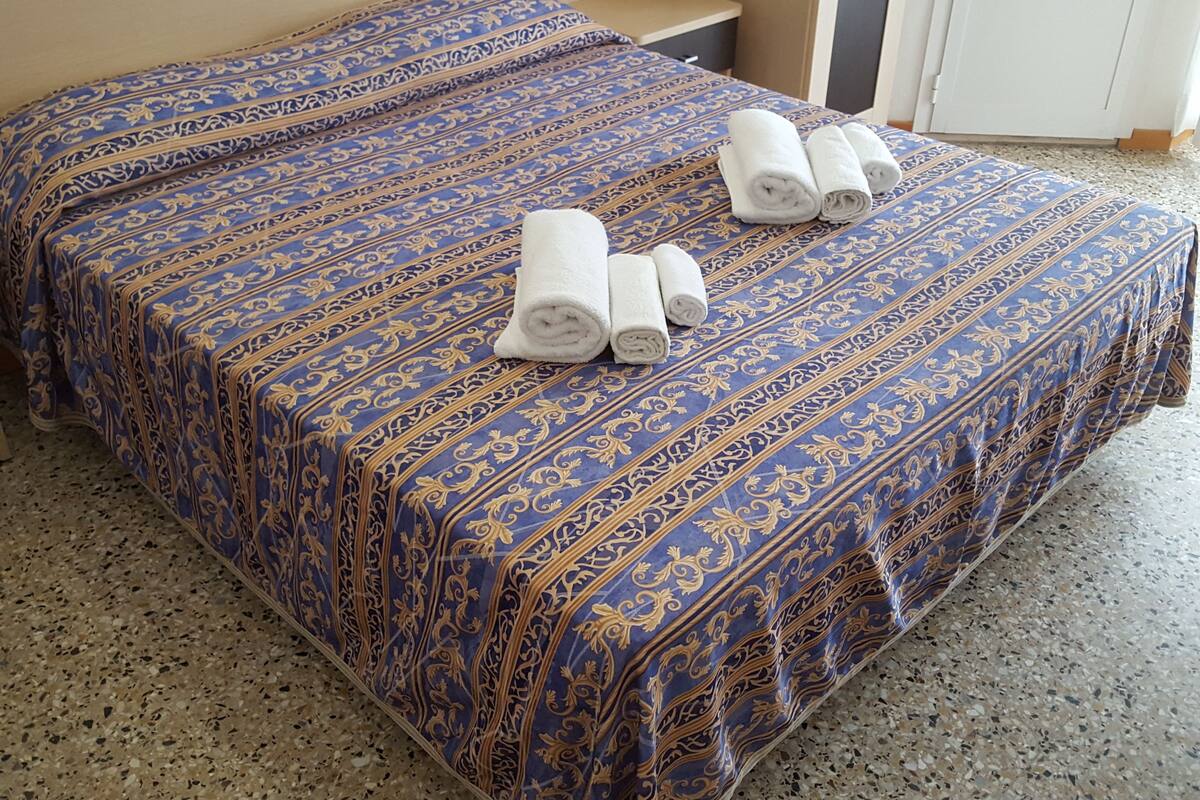 A hotel room with two beds, each covered with a blue and gold patterned bedspread. There are three rolled white towels placed on the beds.