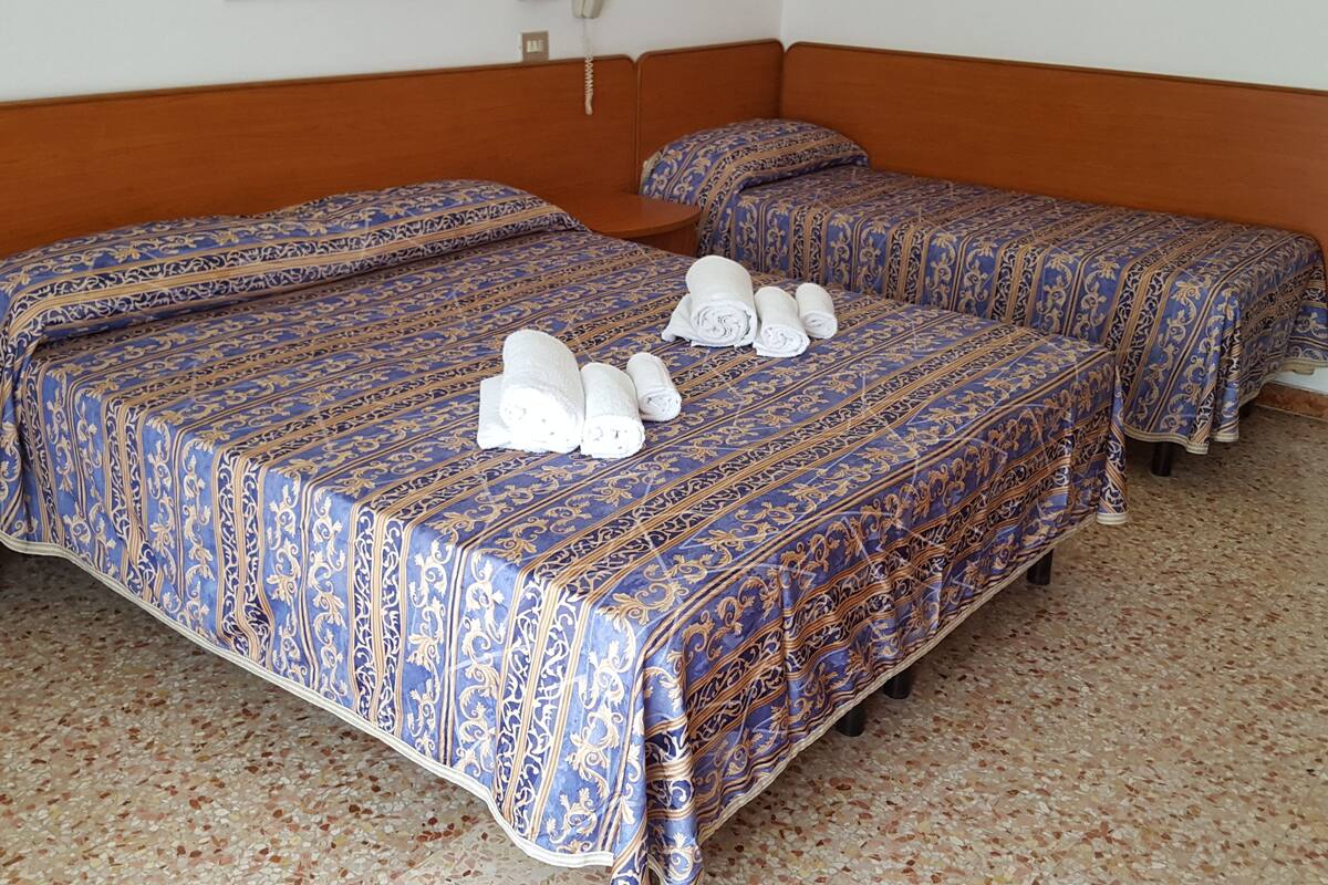 A hotel room with two beds, each with a patterned blue and gold bedspread, and a pair of rolled towels on the bed.