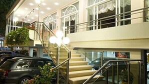 Un hotel con una escalera que lleva a la entrada, un estacionamiento con coches, y una fachada de edificio con amplias ventanas y un balcón.