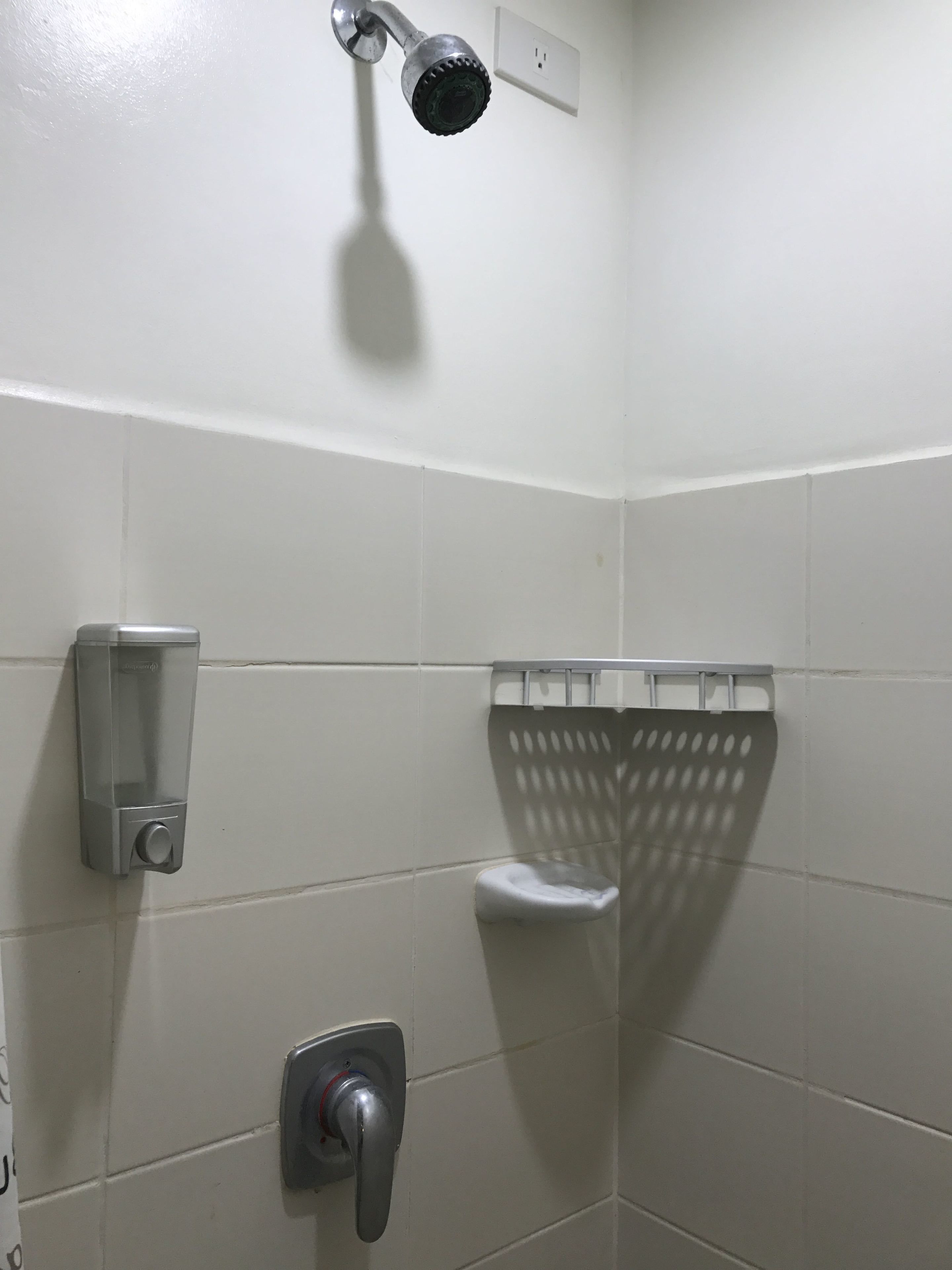Un baño con paredes de azulejos blancos, cabezal de ducha, dispensador de jabón, toallero y porta papel higiénico.