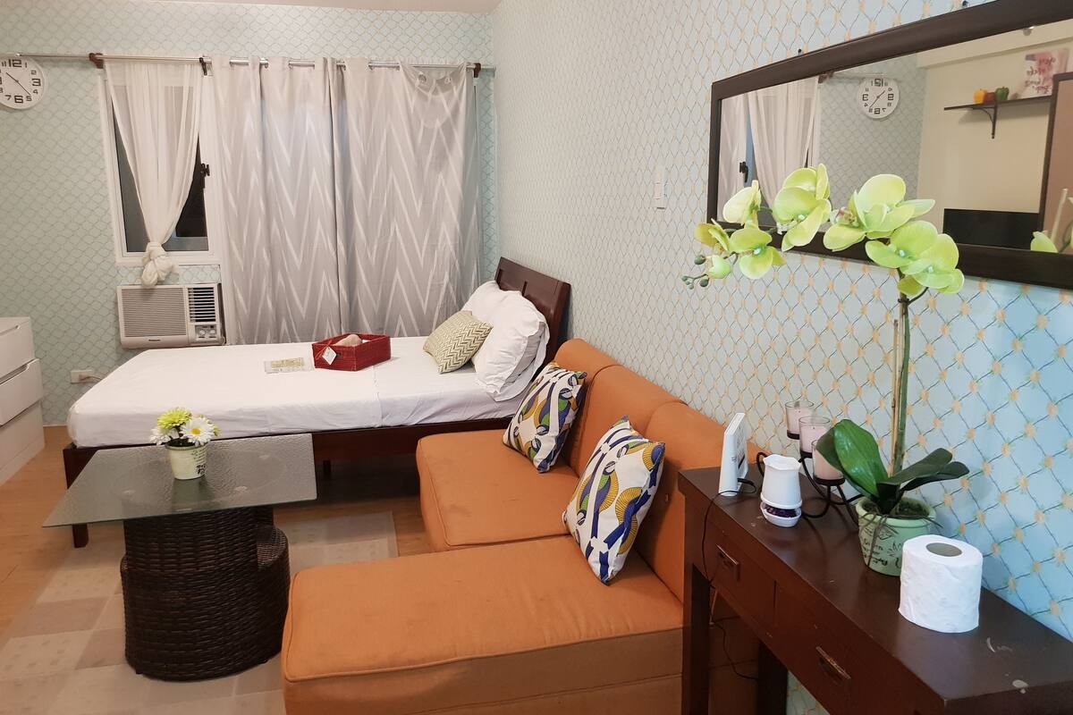 A compact hotel room with a bed, a sofa, a small table with a vase of flowers, a mirror, and a dresser.