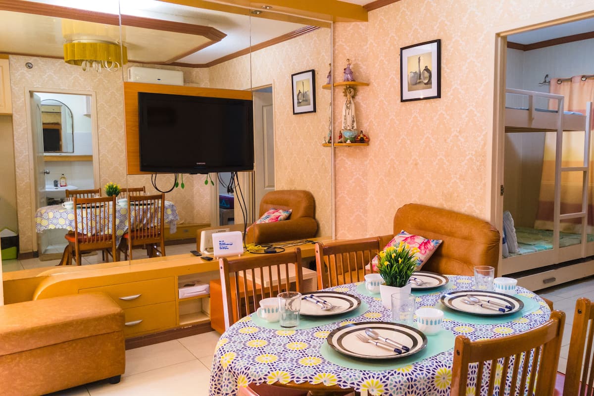 A dining area with a round table set for four, a television above the table, and a sofa in the background.