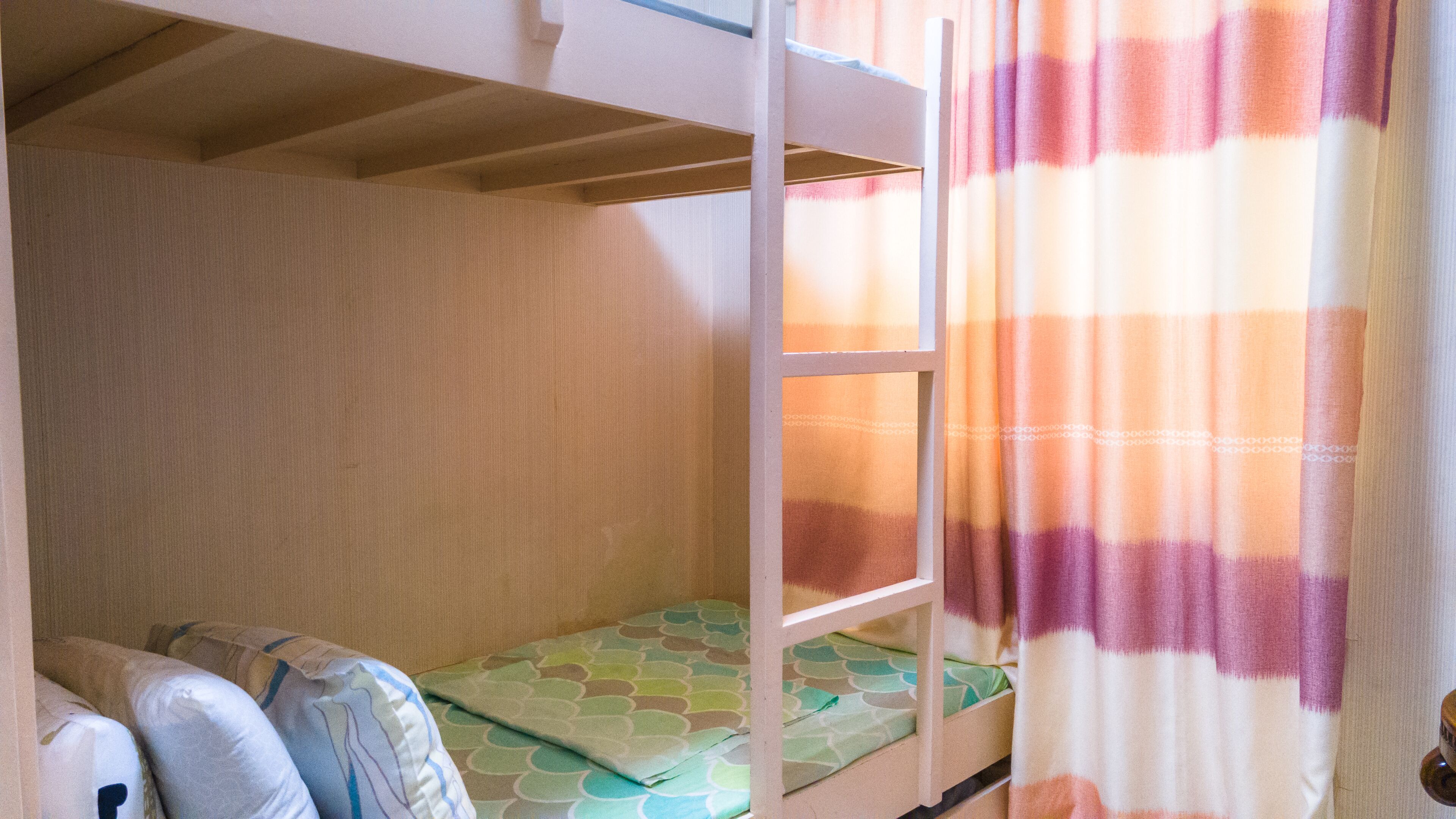 Une chambre avec un lit superposé, un matelas à motifs, une structure en bois et une fenêtre ornée d’un rideau à rayures.