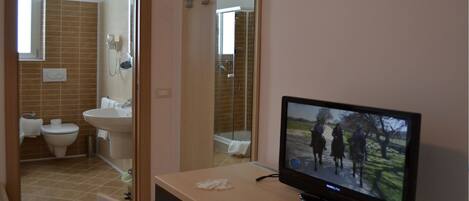 A bathroom with a toilet, sink, and shower. A television is on a stand in the room.