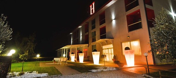 Ein modernes Hotel mit überdachter Eingangspartei, Außenbestuhlung und beleuchteten Pflanzgefäßen.