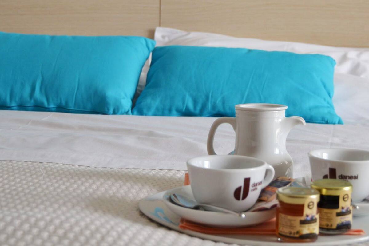 Ein ordentlich bezogenes Bett mit weißer und blauer Bettwäsche, ein Tablett mit zwei weißen Tassen, einer weißen Karaffe und zwei kleinen Teebehältern.