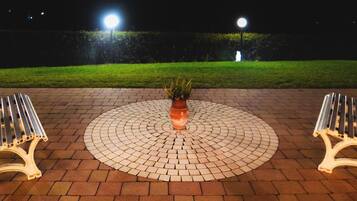 A circular paved area with two white benches on either side, a central potted plant, and a well-lit garden in the background.