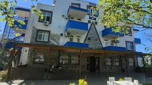 Un edificio de dos plantas con fachada de piedra, un balcón azul y un pequeño espacio al aire libre con sillas blancas.