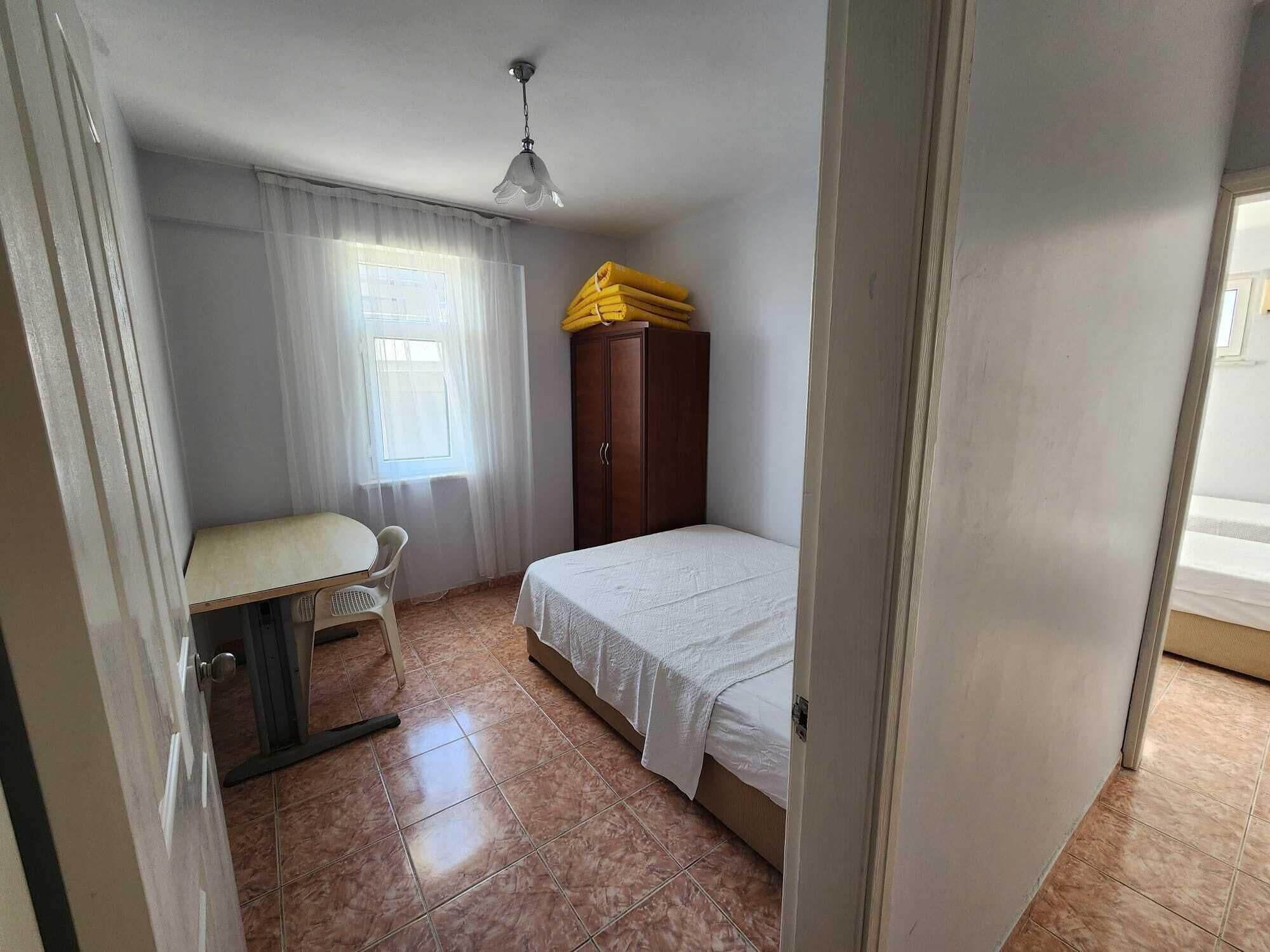 Ein kleines Zimmer mit einem Bett, einem Schreibtisch mit Stuhl, einem Kleiderschrank und einem Fenster mit Vorhängen.