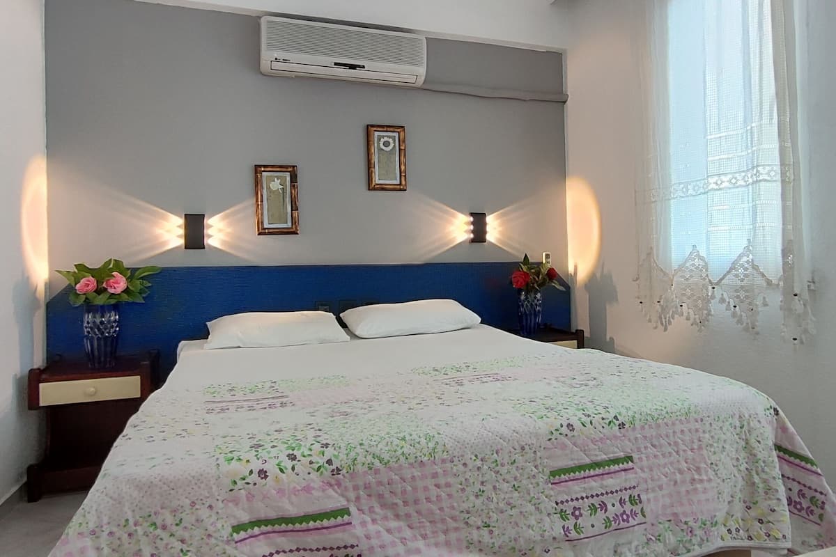 A bedroom with a bed, two wall-mounted lamps, a window with lace curtains, and a wall-mounted air conditioner.