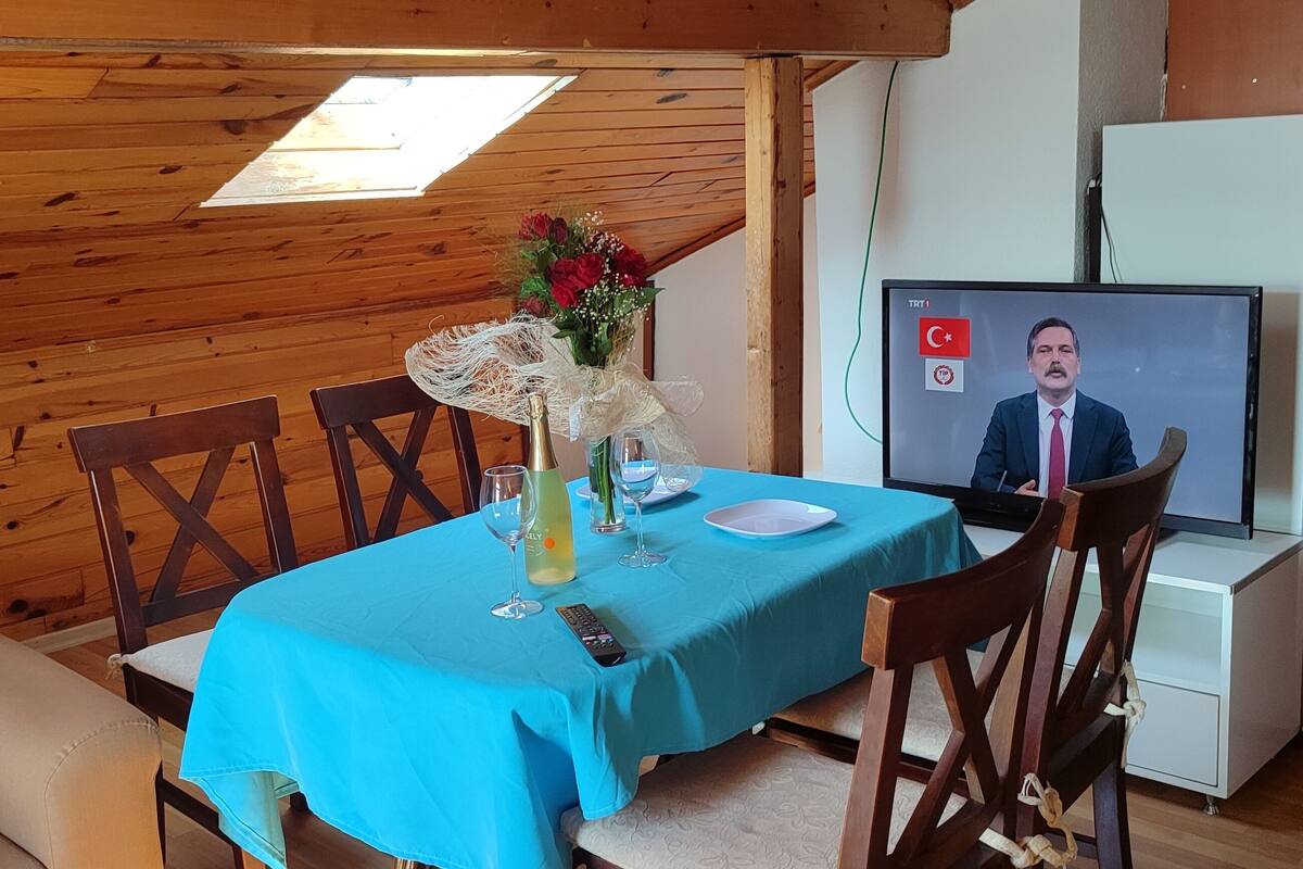 A dining area with a table set for two, a television displaying a person, and a kitchen area with appliances.