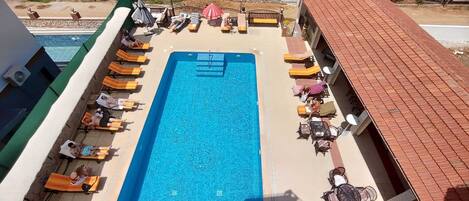 Una piscina rectangular con superficie de agua azul, rodeada de sillas de descanso naranjas y algunos paraguas.
