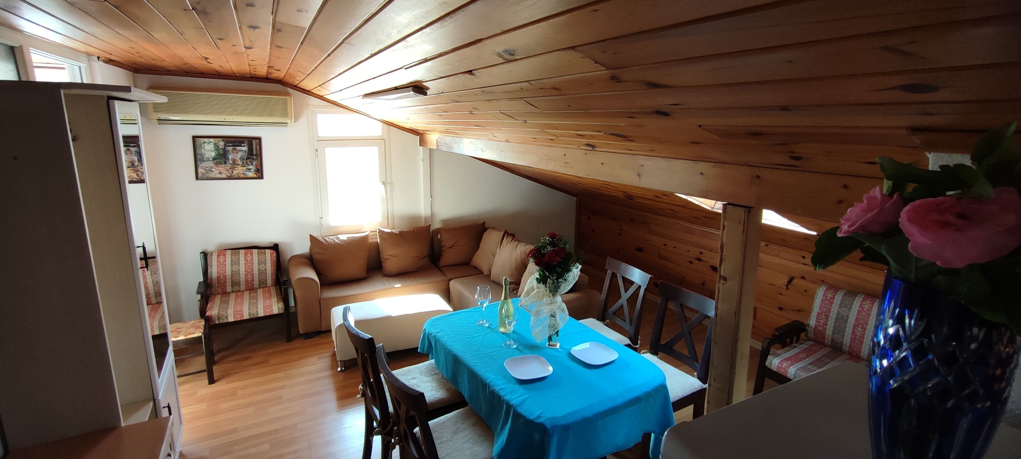 A cozy dining area with a wooden ceiling, a dining table set with plates and a vase of flowers, and a sofa with cushions in the background.