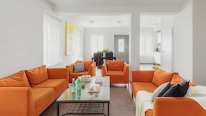 A modern living room with two orange sofas, a glass coffee table, and a dining area in the background. - Swiss Star Oerlikon Lodge (Zürich)