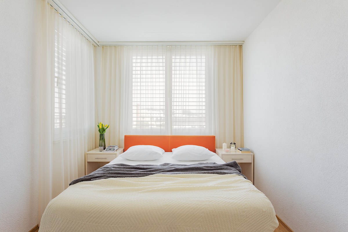 A neatly arranged hotel room with a bed, a nightstand, a window with sheer curtains, and a vase with flowers.