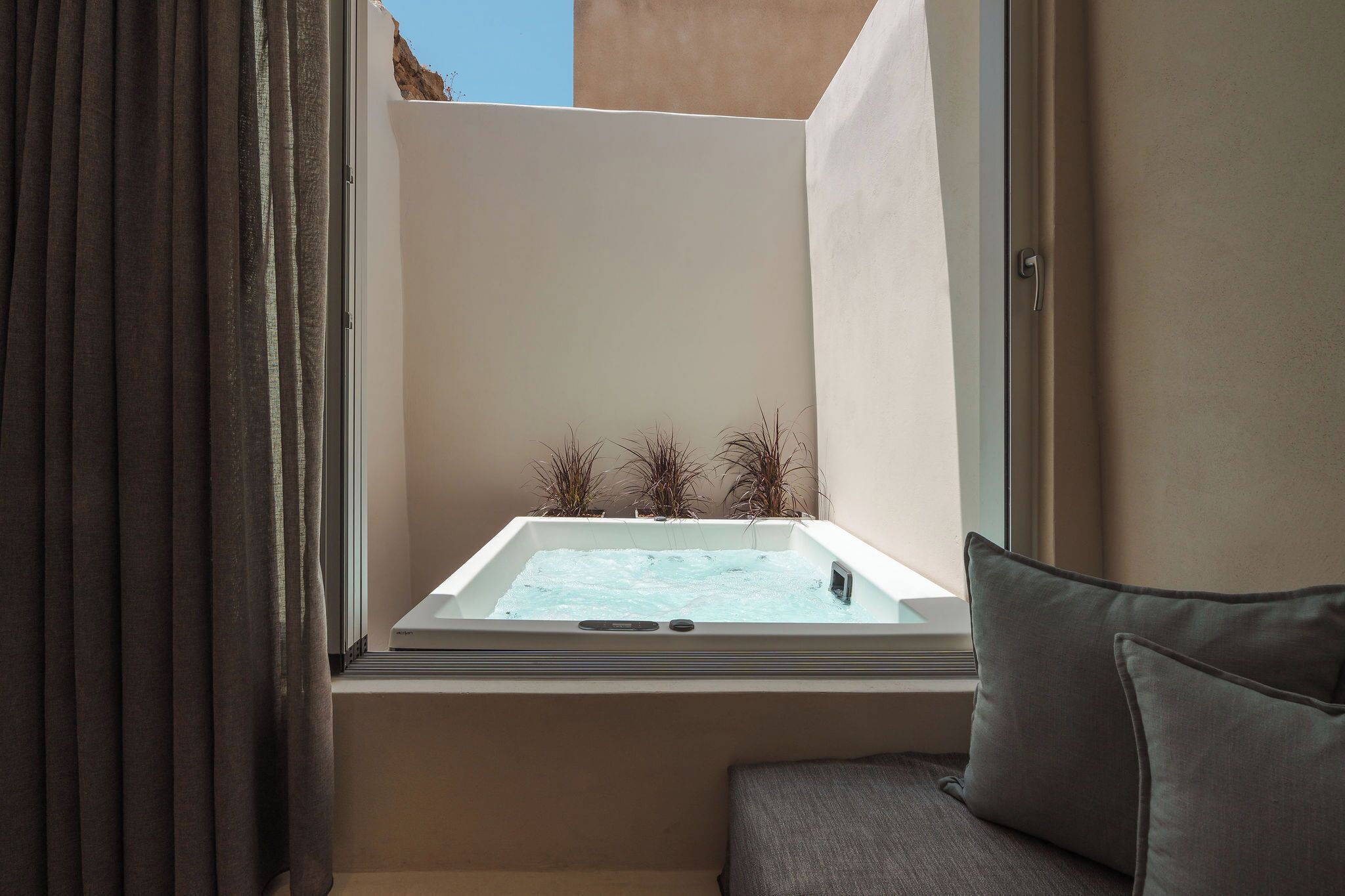 A room with a window overlooking a hot tub, a small plant arrangement, and a grey sofa.