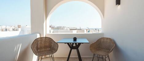 A balcony with a table and two wicker chairs, a view of the city through an arched window, and a wall-mounted light fixture.