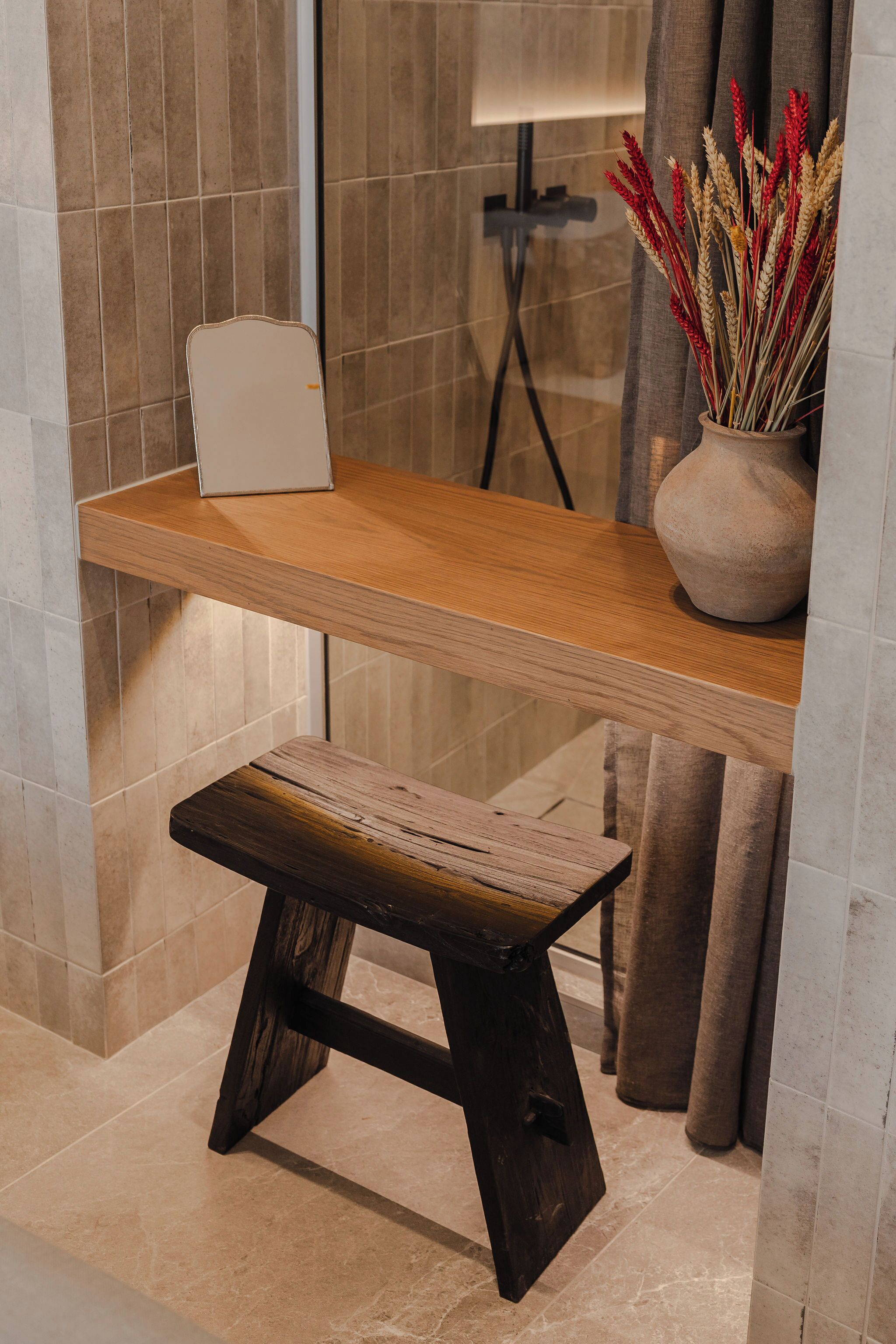 A wooden desk with a laptop, a vase with dried plants, and a bench in a modern bathroom with tiled walls and floor.