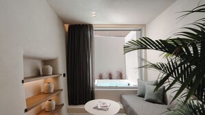 Une pièce moderne et compacte, avec un canapé gris, une table basse ronde et des étagères ornées d’objets décoratifs. Une grande fenêtre offre une vue sur une piscine.