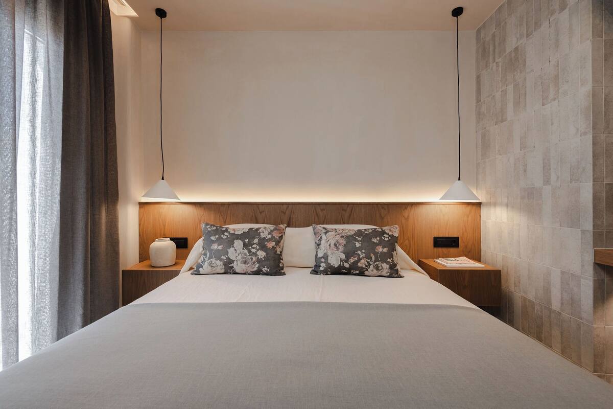 Una cama bien hecha con dos almohadas, una mesita de noche con lámpara y una pared con textura.