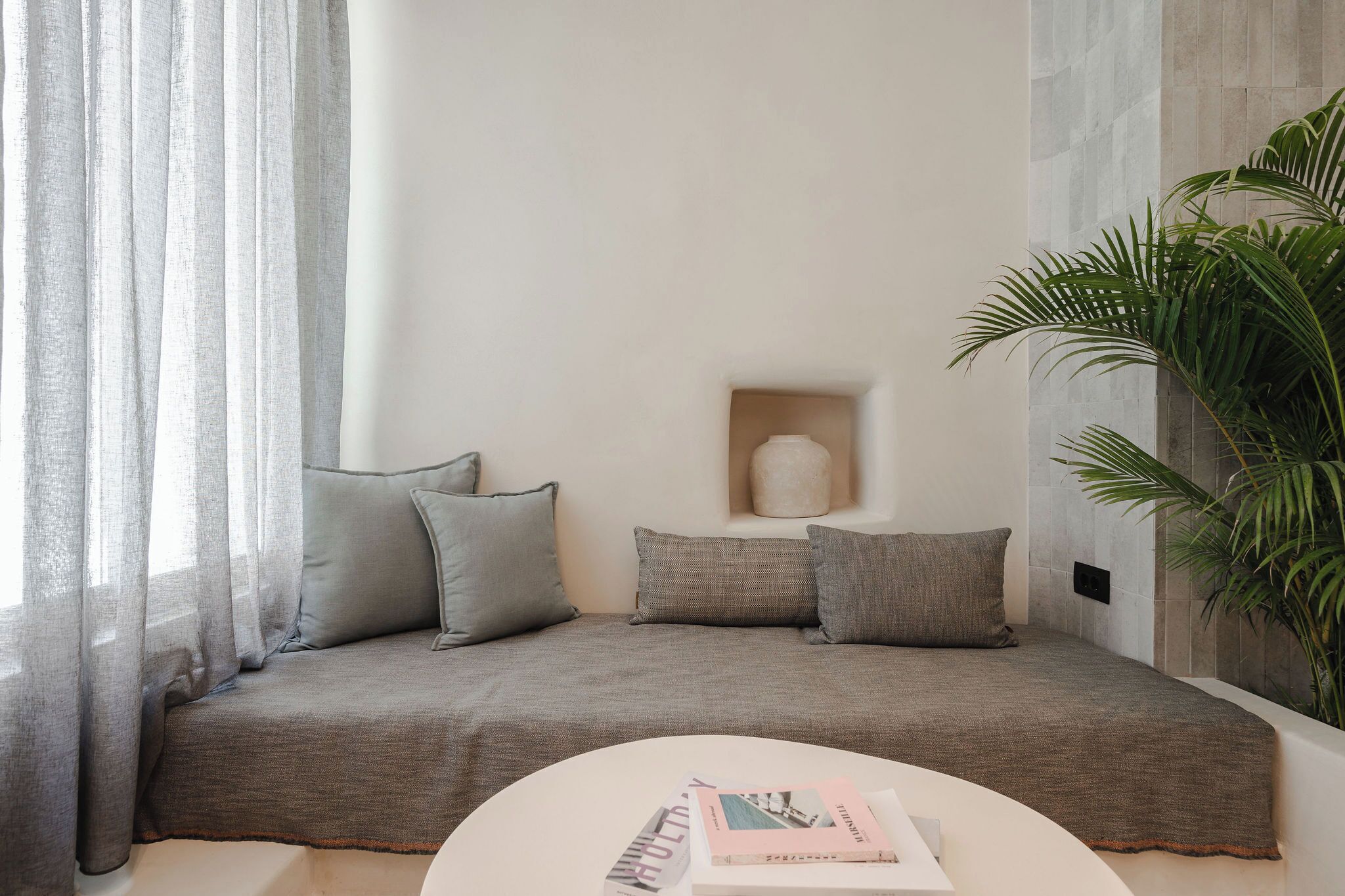 A modern living room with a sofa, pillows, a small table with books, a plant, and a wall-mounted shelf.