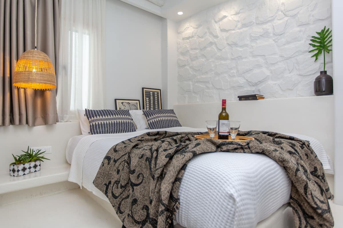 Un dormitorio moderno con cama, lámpara colgante, pared con textura de piedra, una planta pequeña y una botella de vino sobre una bandeja.
