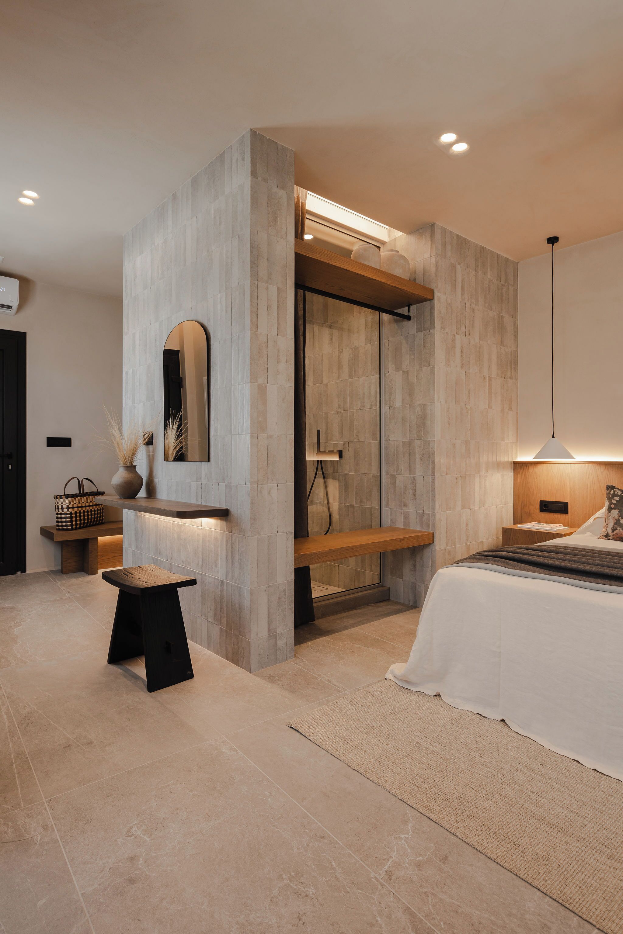 A modern bedroom with a large bed, a wooden bench, a small table, a mirror, and a built-in shower.