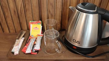 Uma chaleira elétrica de aço inoxidável, uma caneca de vidro e sachês de café instantâneo sobre uma superfície de madeira.