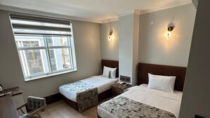Une chambre d’hôtel avec deux lits simples, un bureau, une chaise, une fenêtre avec stores et un luminaire au plafond.