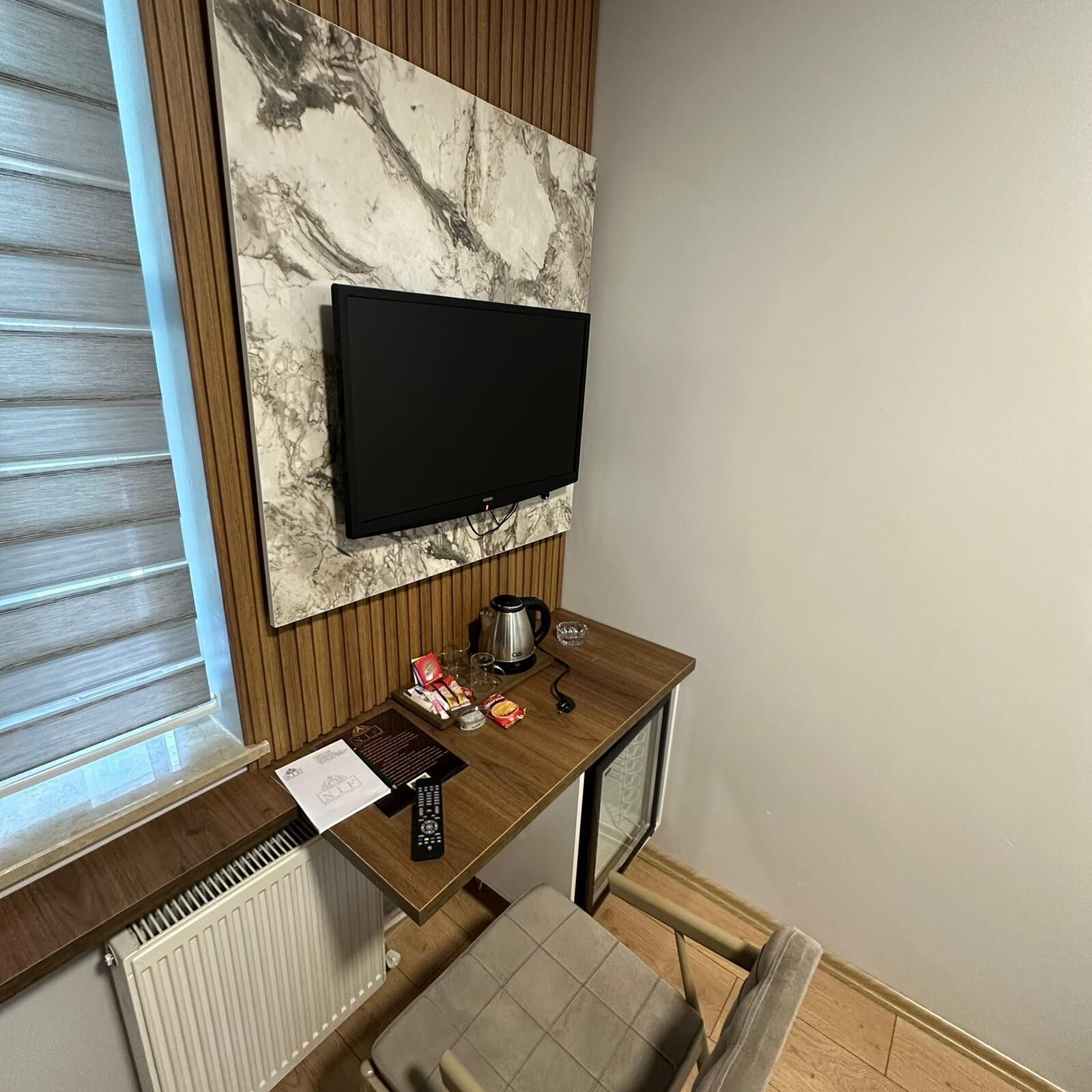 A small room with a television mounted on the wall, a wooden desk with a kettle, a cup, and some snacks, a radiator, and a window with blinds.