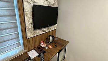 A small room with a television mounted on the wall, a wooden desk with a kettle, a cup, and some snacks, a radiator, and a window with blinds.