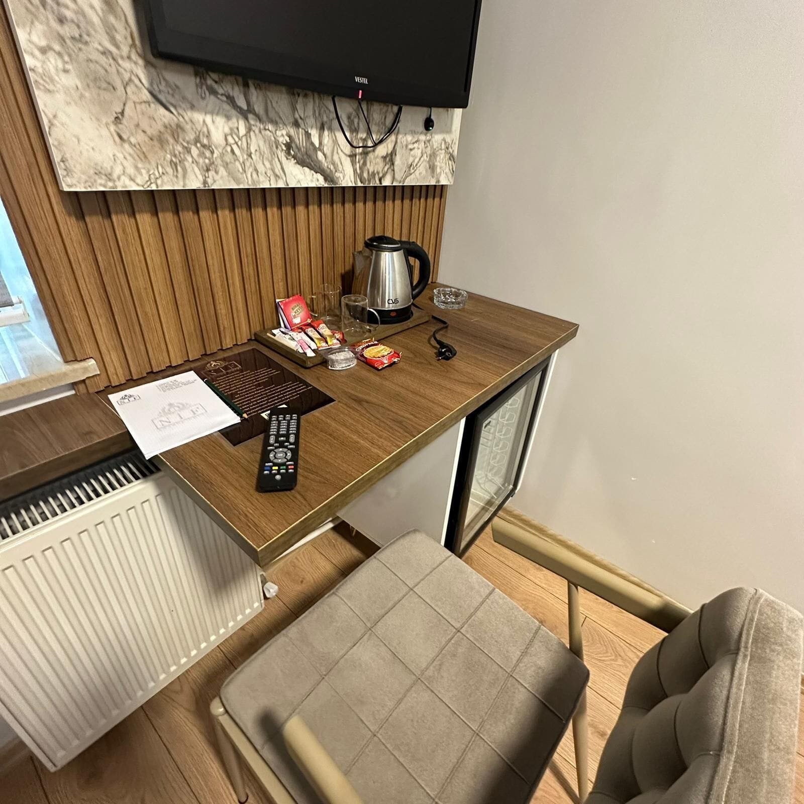 A compact hotel room with a wooden desk, a television mounted above it, a chair, a radiator, and a small kitchenette area.
