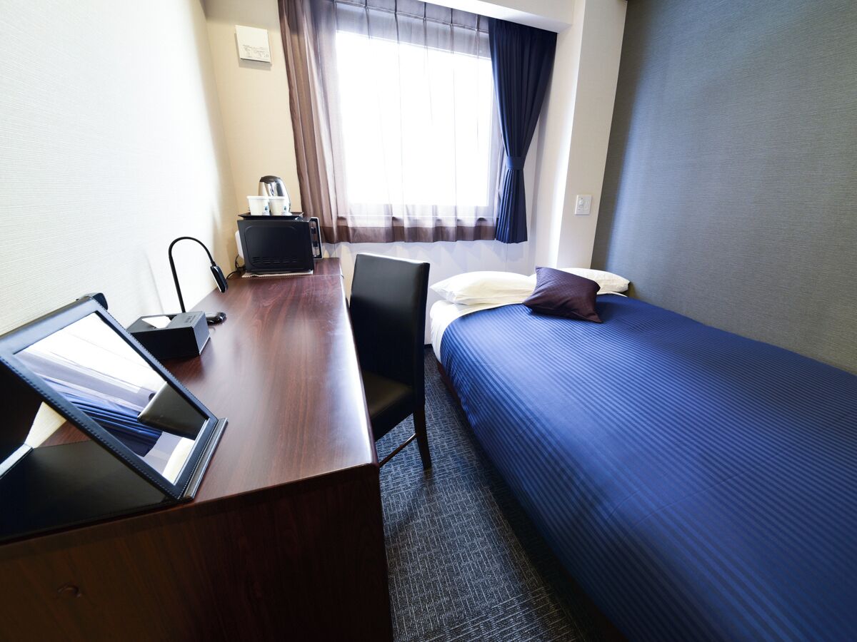 A hotel room with a wooden desk, a laptop, a bed with blue bedding, a chair, and a window with curtains.