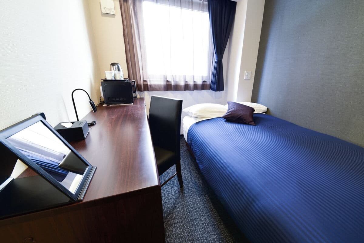 A hotel room with a wooden desk, a laptop, a bed with blue bedding, a chair, and a window with curtains.