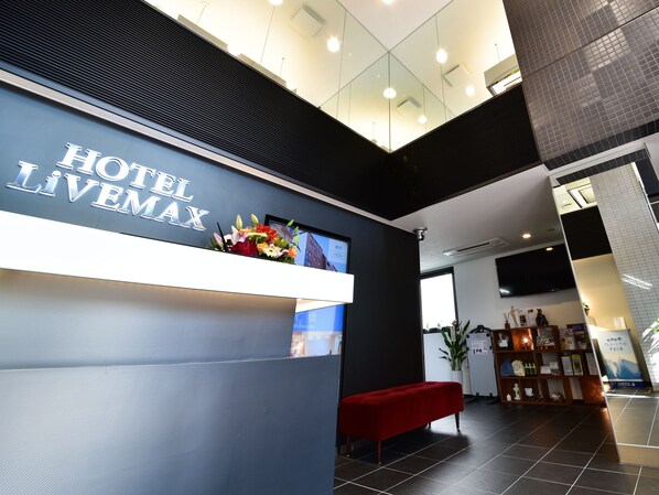 A hotel reception desk with a sign reading "HOTEL LIVEMAX". There is a red bench, a potted plant, and a shelf with various items including a framed picture and a small statue.