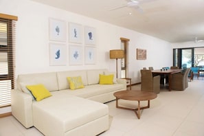 Uma sala de estar moderna com um sofá bege, almofadas amarelas, uma mesa de centro de madeira e uma área de jantar com cadeiras de vime.