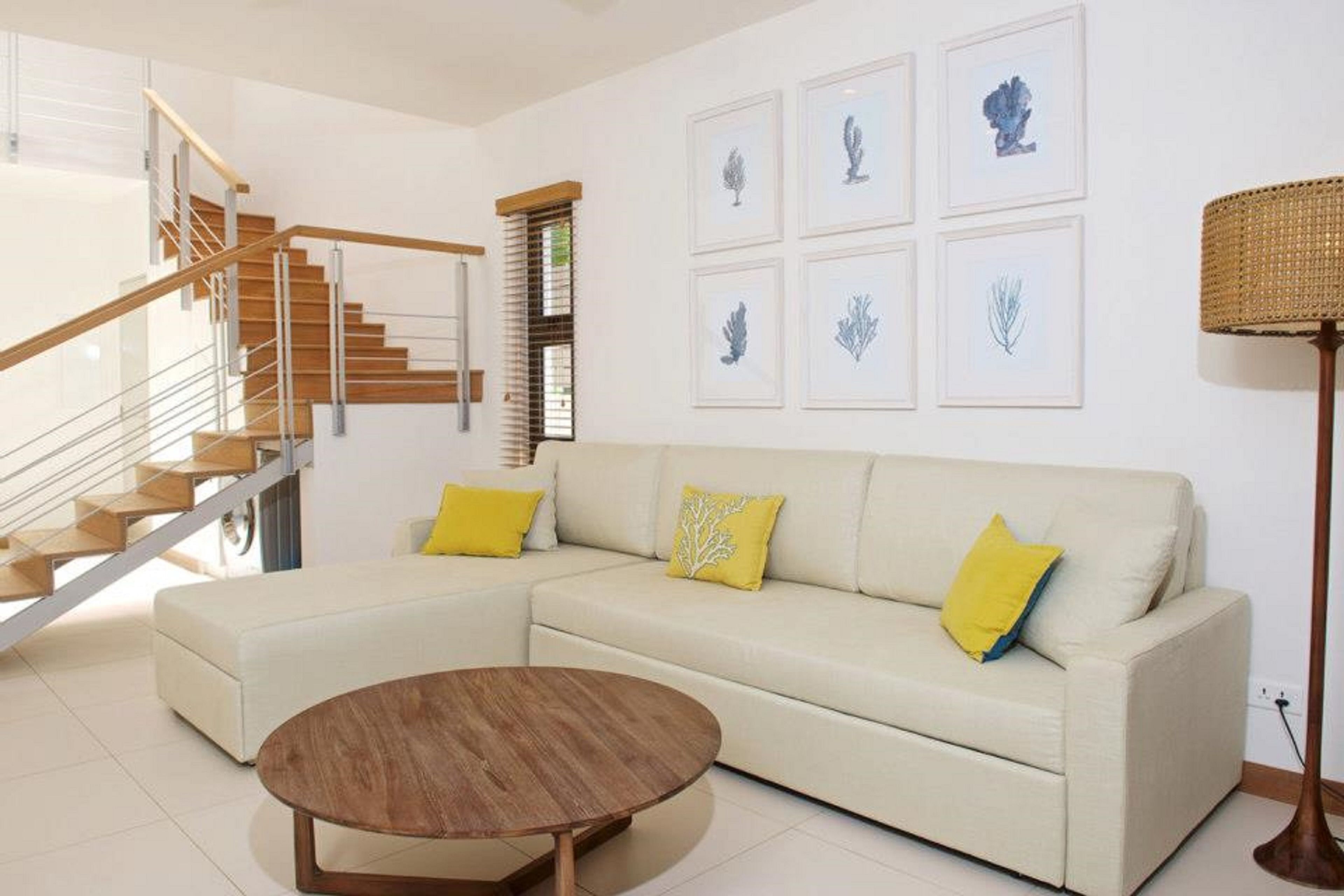 Una sala moderna con una escalera de madera, un sofá seccional beige con cojines amarillos, una mesa de centro redonda de madera e impresiones botánicas enmarcadas en la pared.