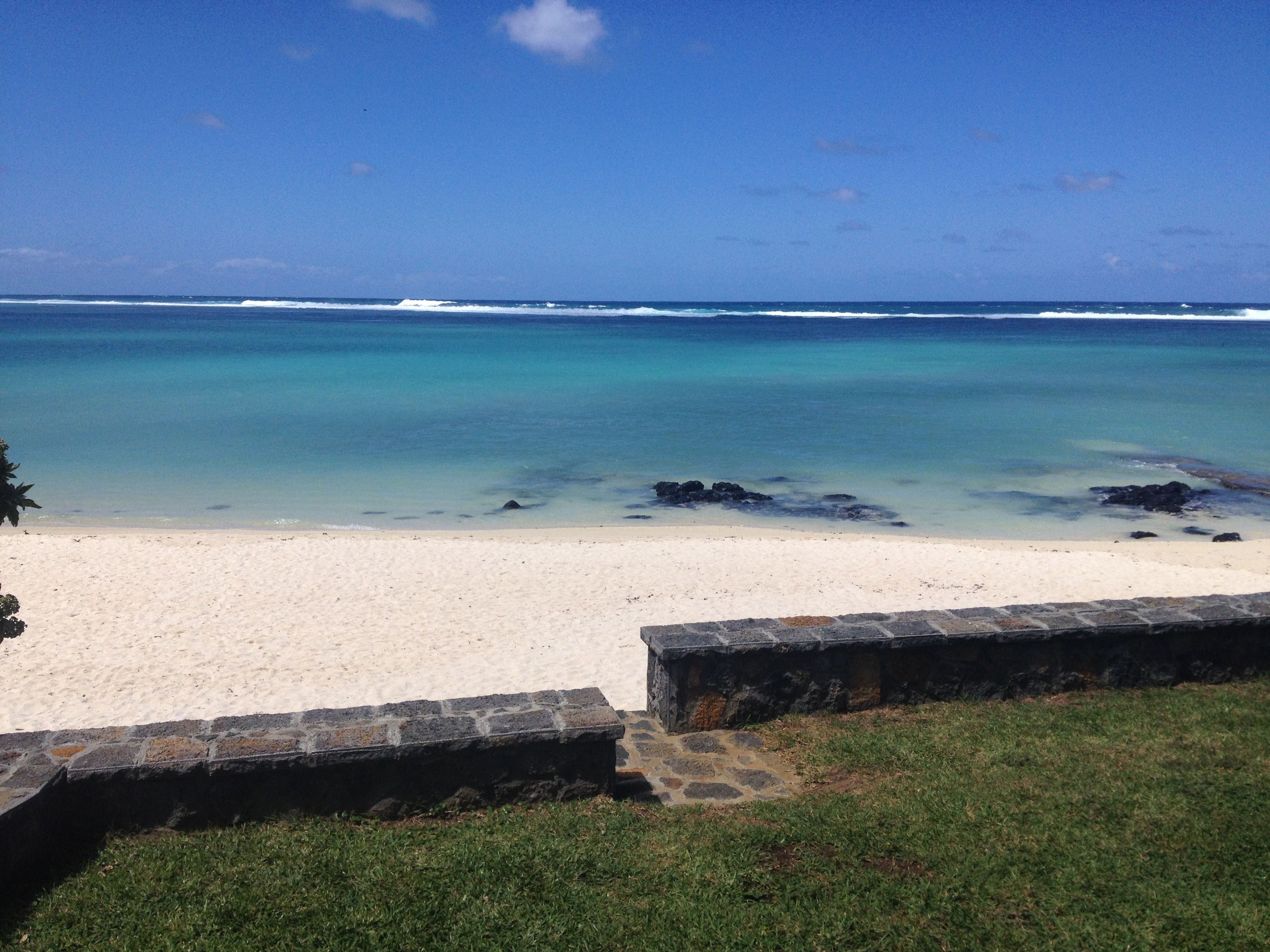 Une plage avec une eau turquoise et limpide, du sable blanc et un mur de pierre au premier plan.