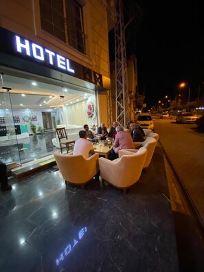 Exterior de un hotel con fachada de vidrio, zona de estar al aire libre y entrada visible.