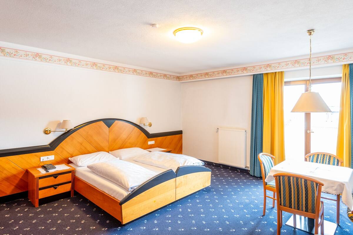 A hotel room with a wooden bed, bedside tables, a desk with chairs, a window with curtains, and a radiator.