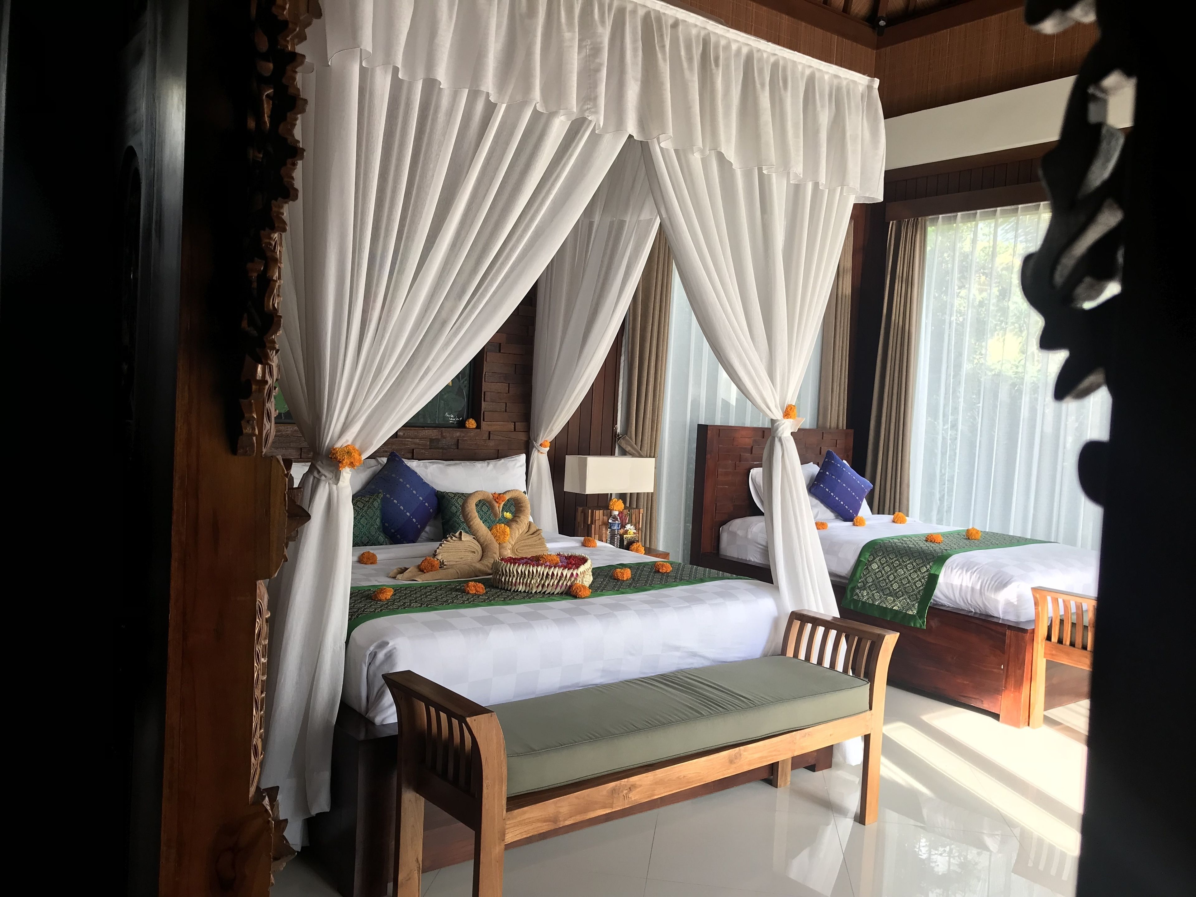 A room with two beds, each with a canopy bedspread and pillows, a wooden bench, and a small table with a lamp.