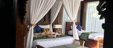 A room with two beds, each with a canopy bedspread and pillows, a wooden bench, and a small table with a lamp.