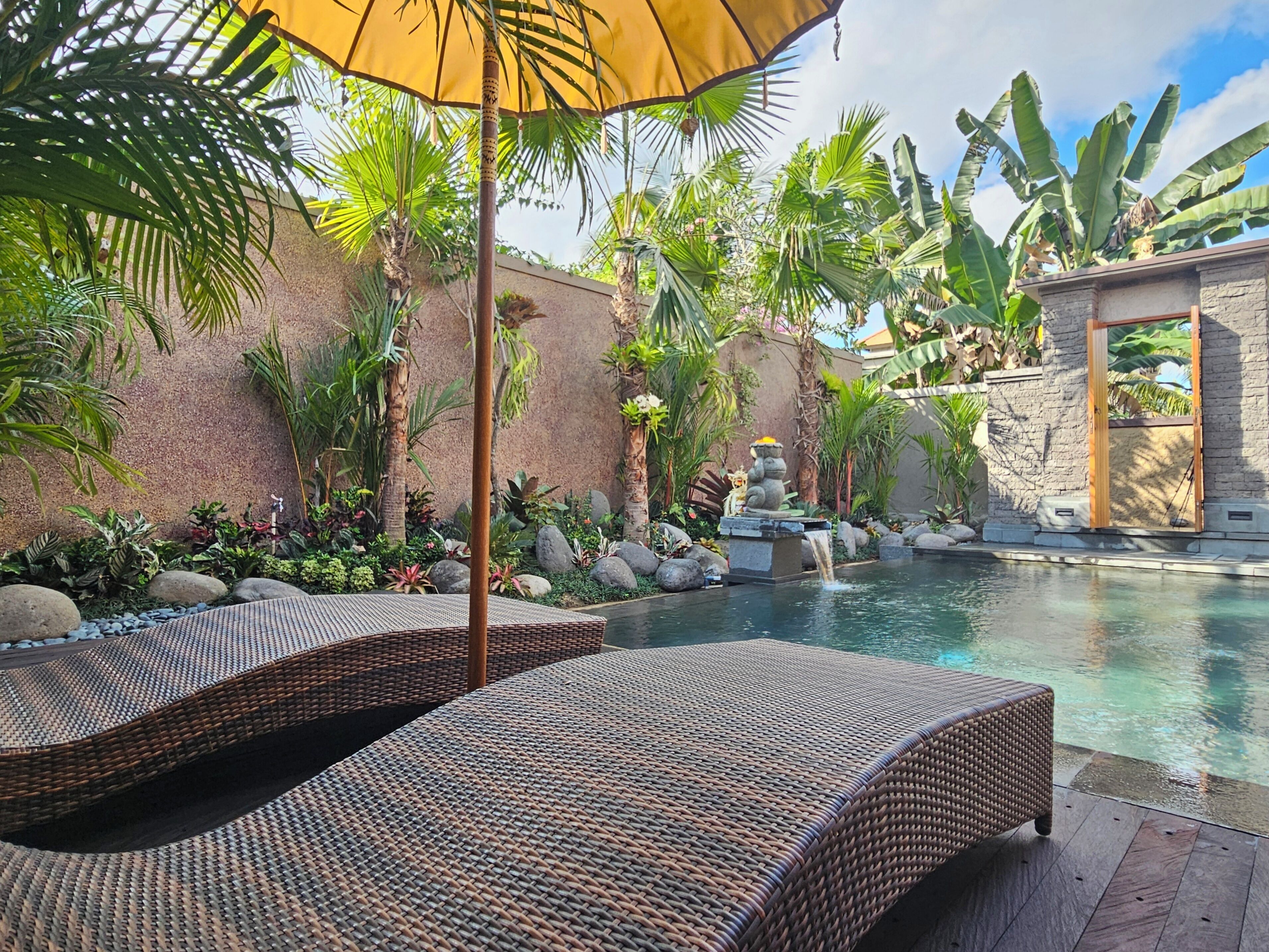 Un área de piscina con sillones de mimbre, una estatua de piedra y plantas tropicales.