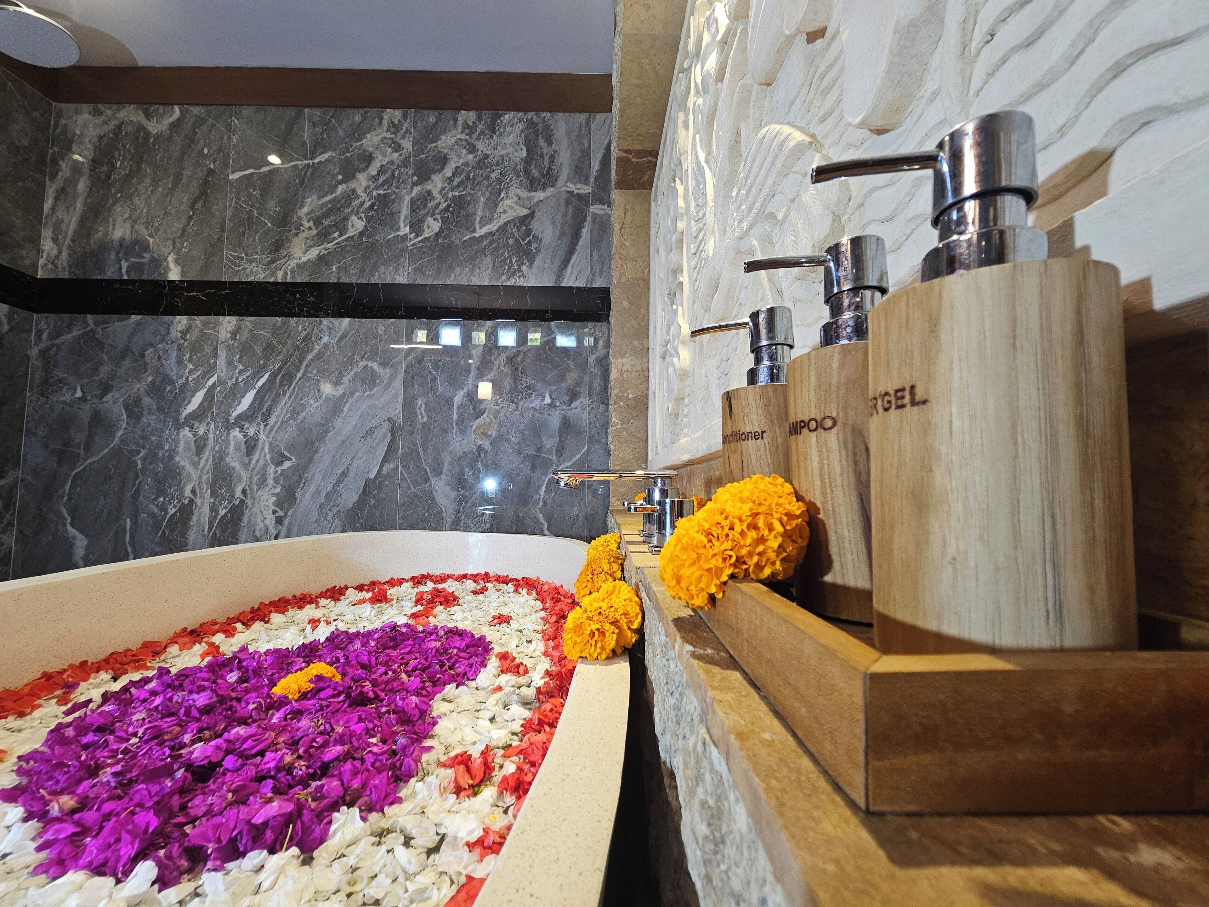 A bathroom with a marble wall, a bathtub filled with flower petals, and wooden shampoo dispensers.