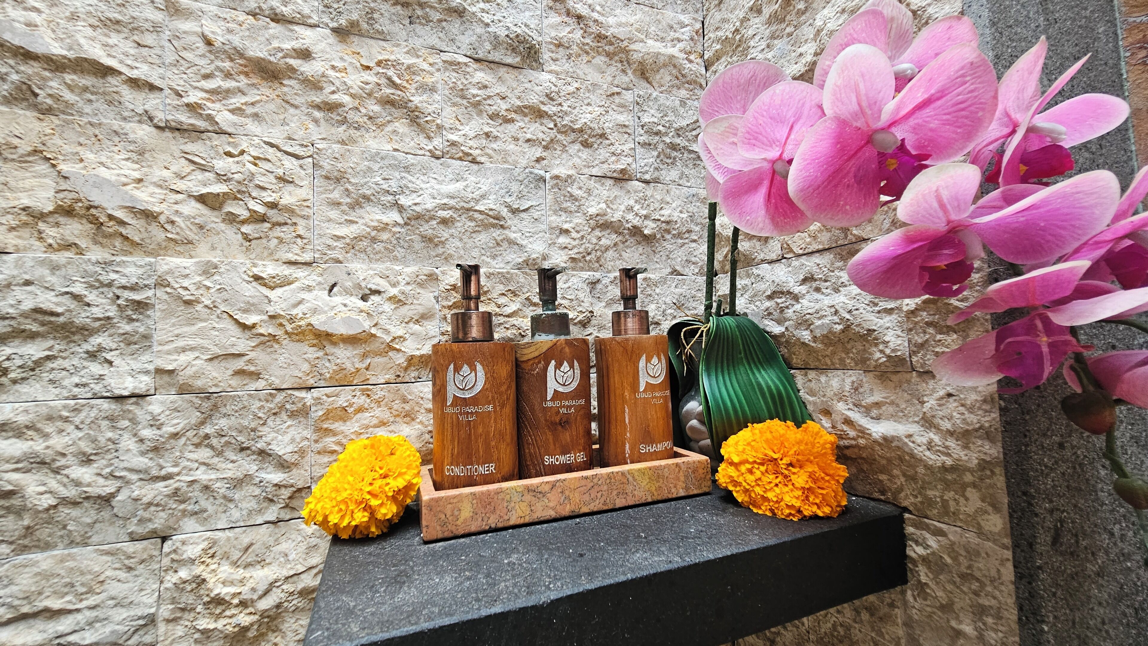 A shelf with four bottles of Ubud Paradise Villa shower gel, a green hairbrush, and two marigold flowers.