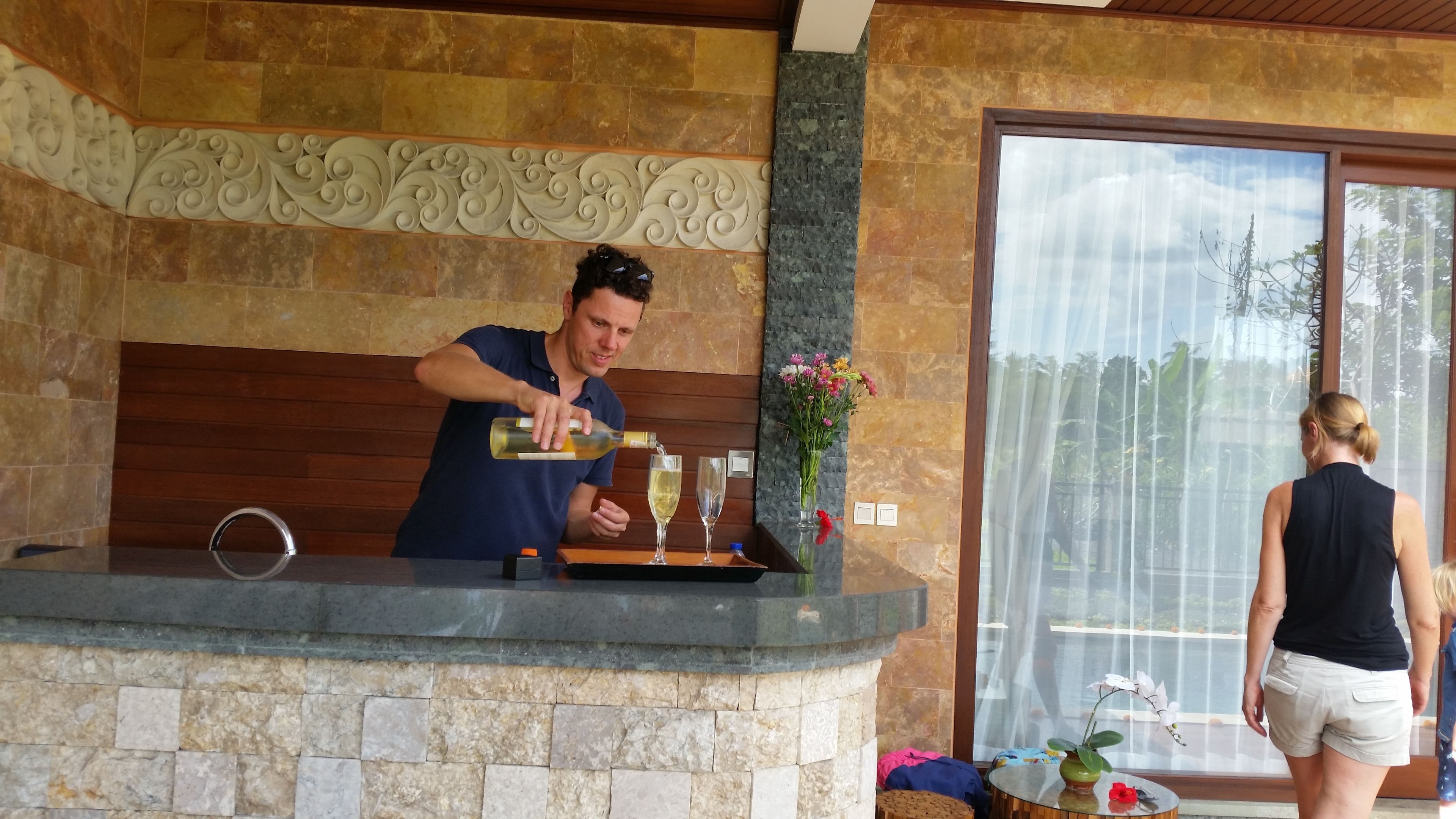 A man pouring a drink into a glass at a counter with stone walls and a wooden panel.