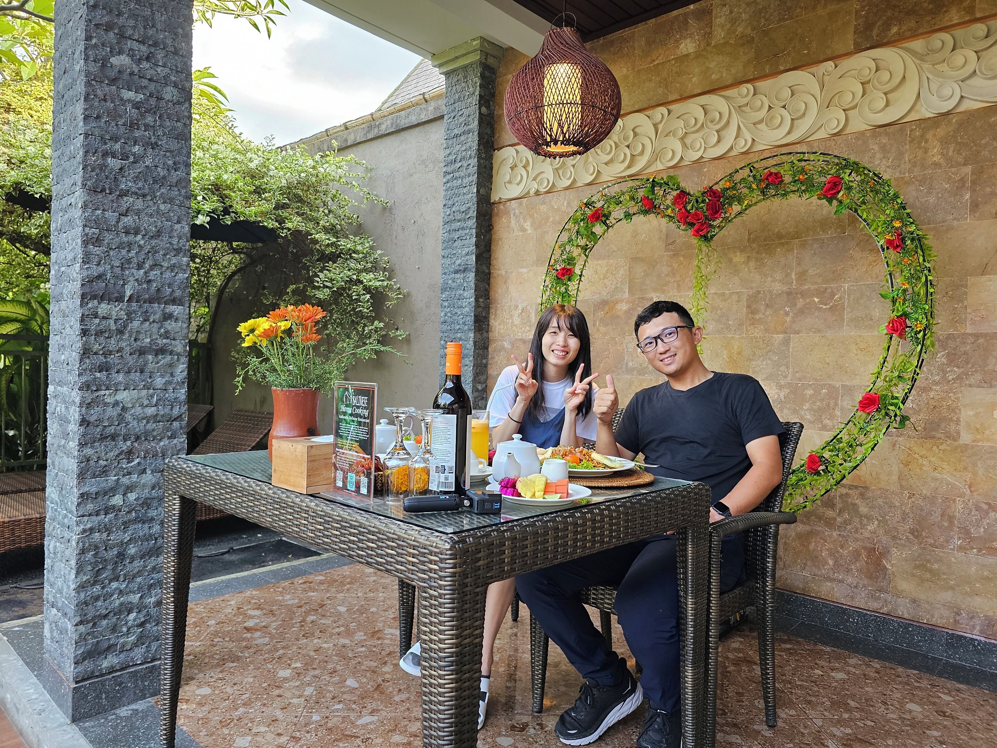 Una pareja cenando en una mesa al aire libre con un arreglo decorativo en forma de corazón y una lámpara colgante.