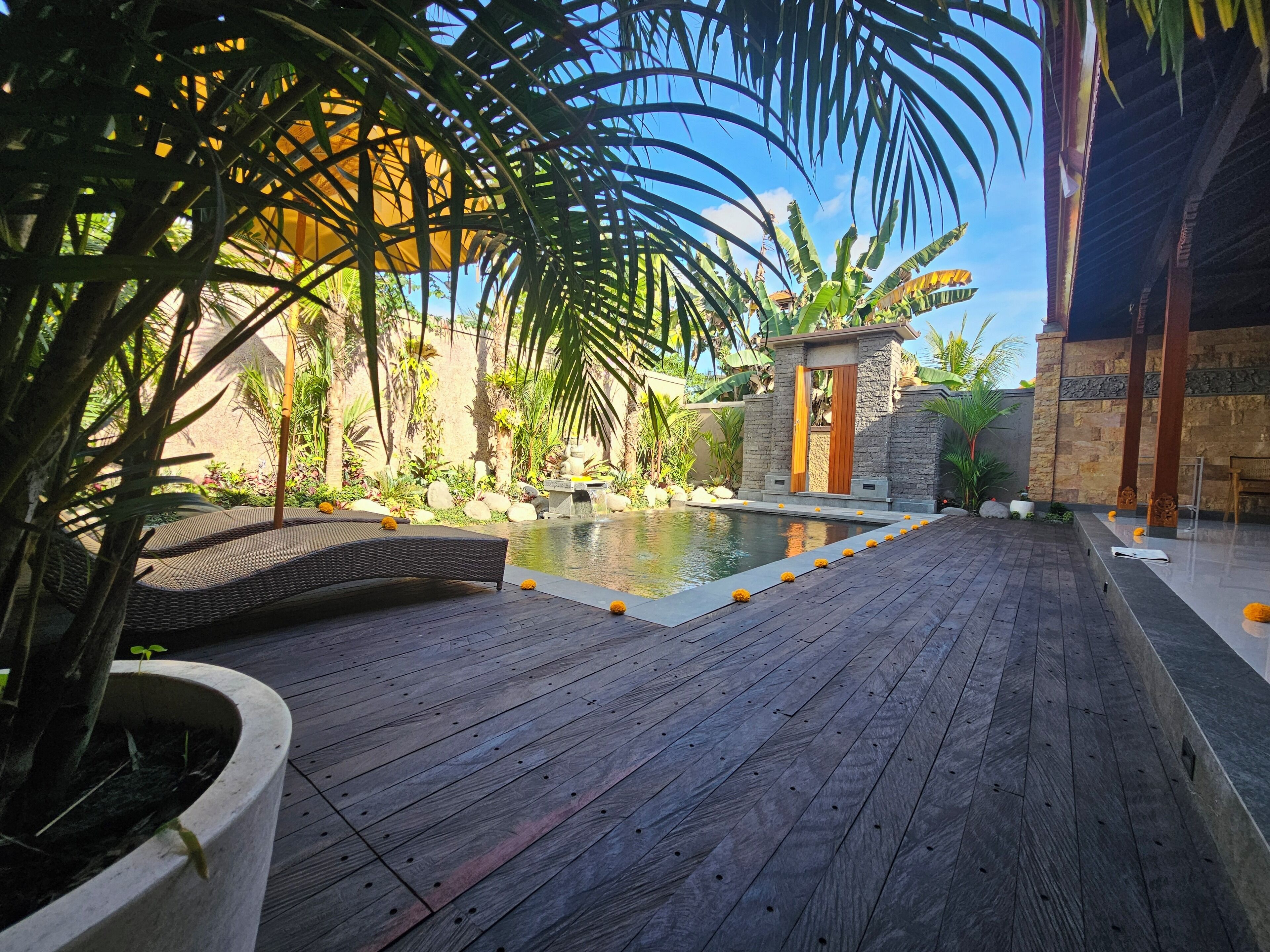 A wooden deck with a pool, surrounded by tropical plants and a stone structure.