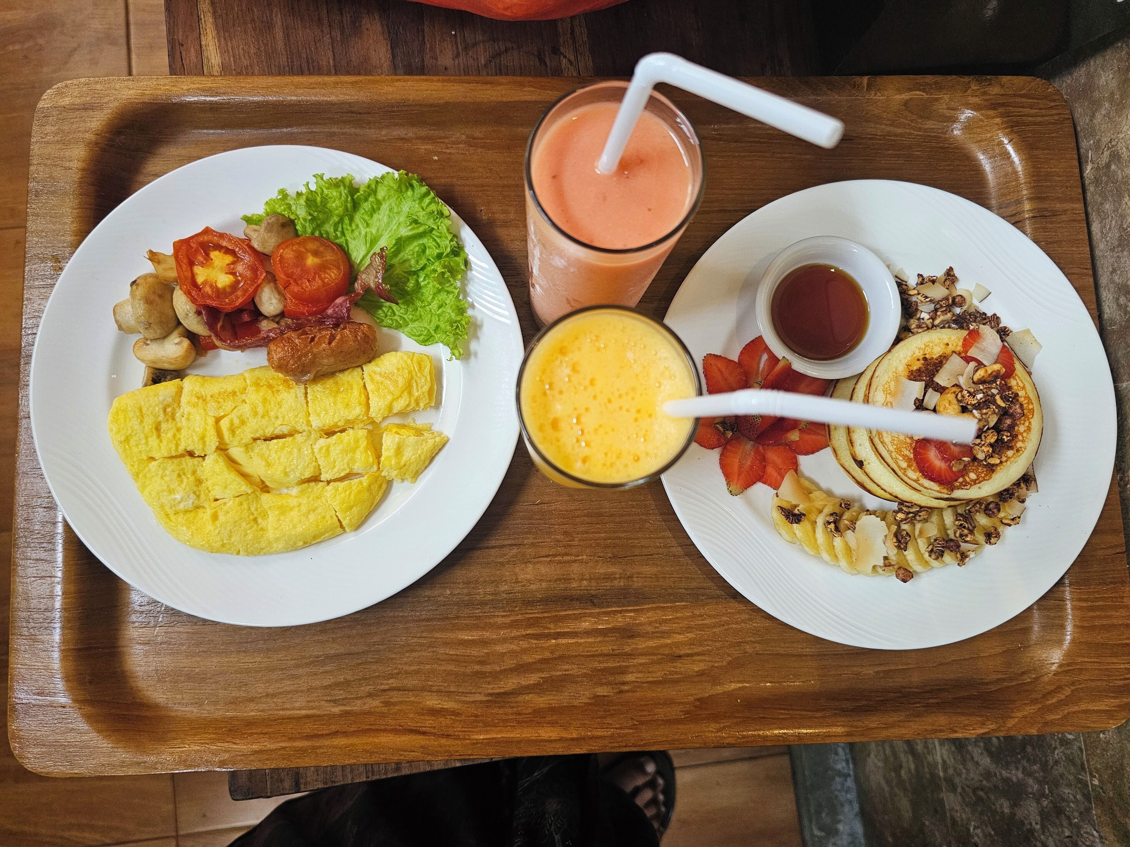 Two plates of food on a wooden tray, including scrambled eggs, bacon, tomatoes, lettuce, and a smoothie with a straw.